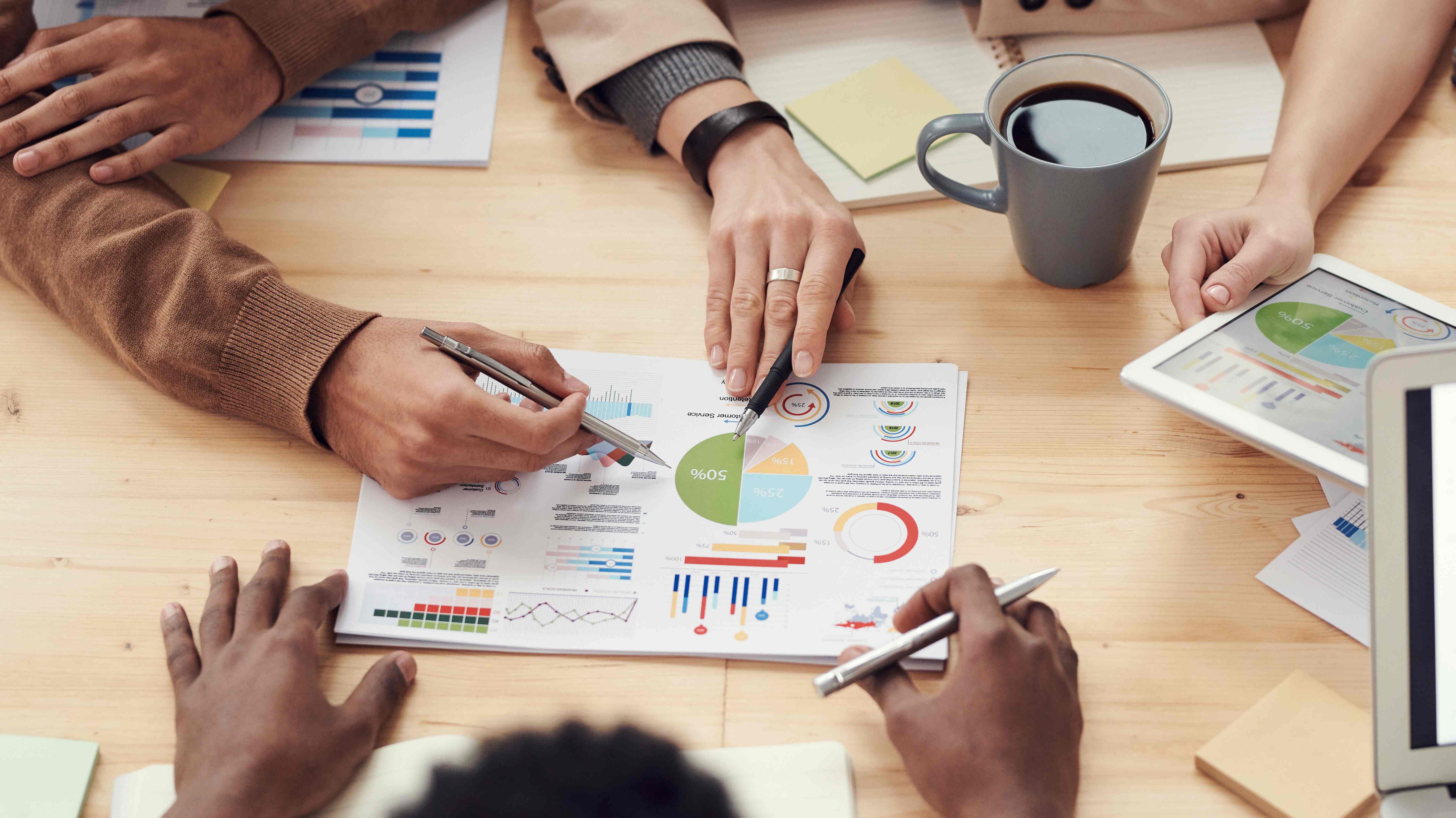 Many people in a meeting pointing at a piece of paper in the center of a table with graphs illustrated on the paper. 3 different sets of hands, each with a pen in their hand.